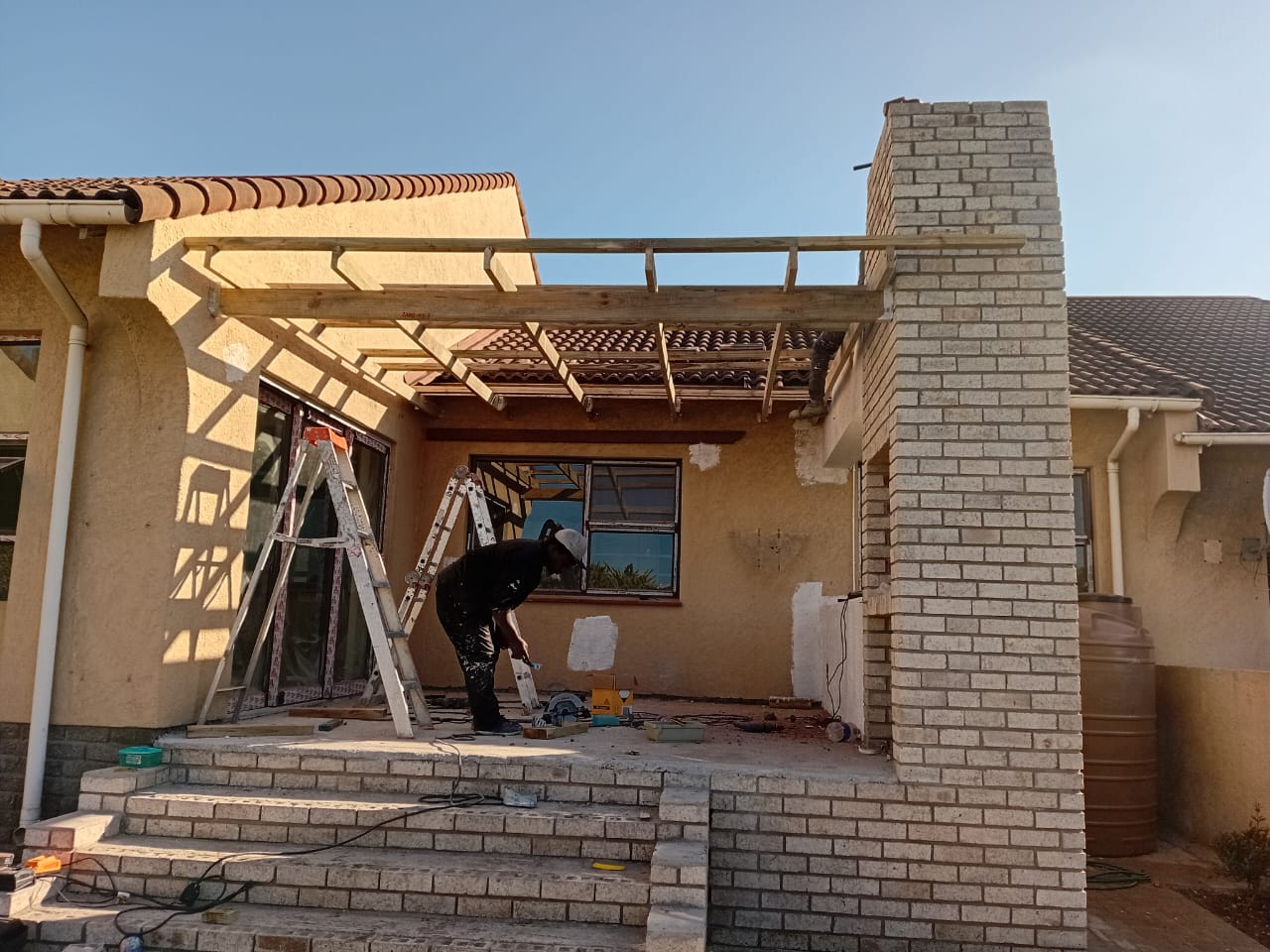 Before: Flat roof installation Port Elizabeth — open patio with bare timber beam frame structure being built, worker drilling, brick column and tile roof behind